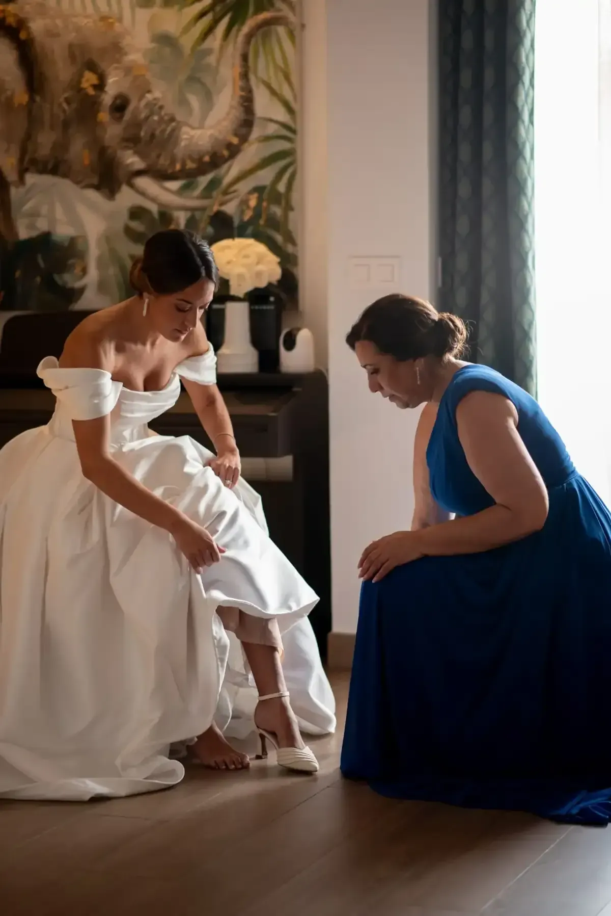 Novia con vestido blanco ajustándose el zapato con la ayuda de una mujer con vestido azul en el interior.
