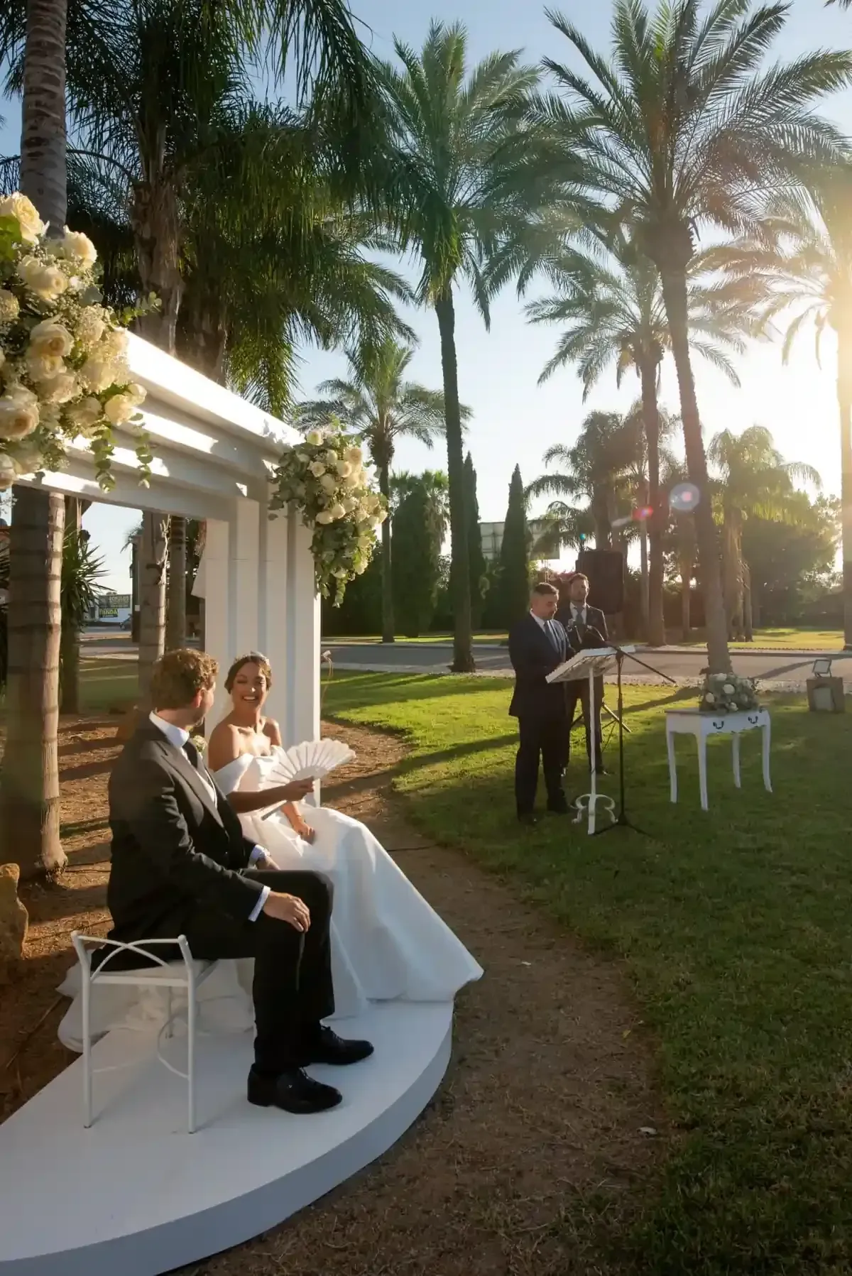 Ceremonia nupcial al aire libre junto a un pabellón blanco con palmeras e invitados sentados bajo la luz del atardecer.