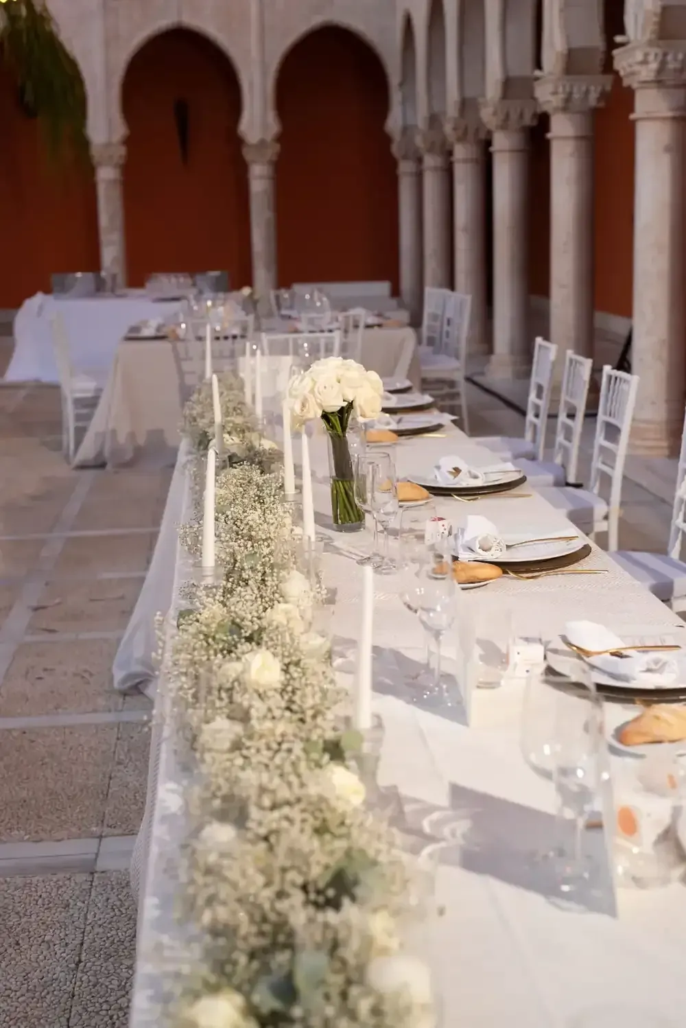 Larga mesa de banquete con sillas blancas y guirnalda floral en un patio de piedra con arcos.