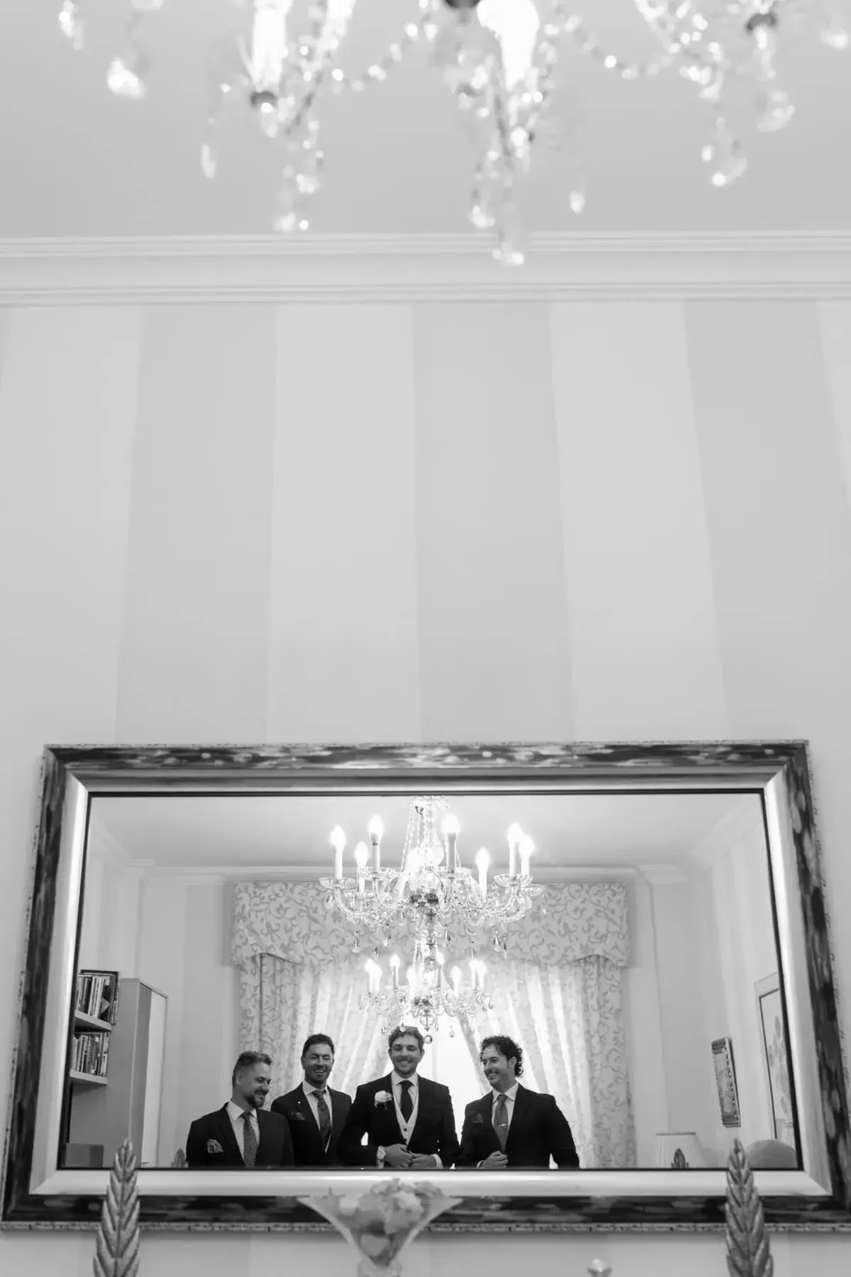 Cuatro personas posando frente a un espejo bajo una lámpara de araña en una habitación luminosa y ornamentada.