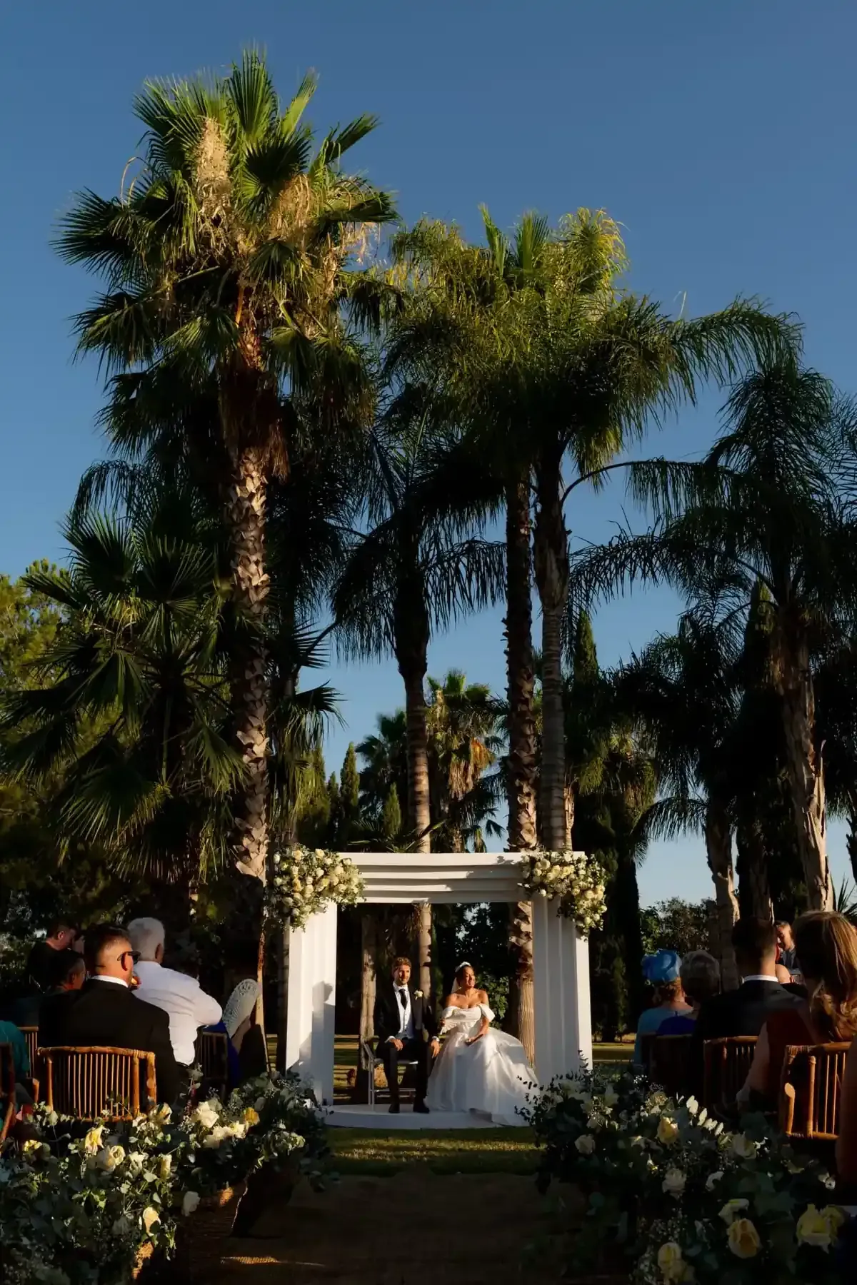 Ceremonia de boda al aire libre bajo altas palmeras con un arco blanco e invitados sentados bajo la cálida luz del sol.