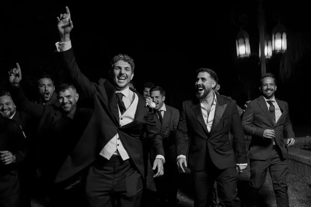 Hombres con traje bailando y vitoreando en una calle poco iluminada por la noche.