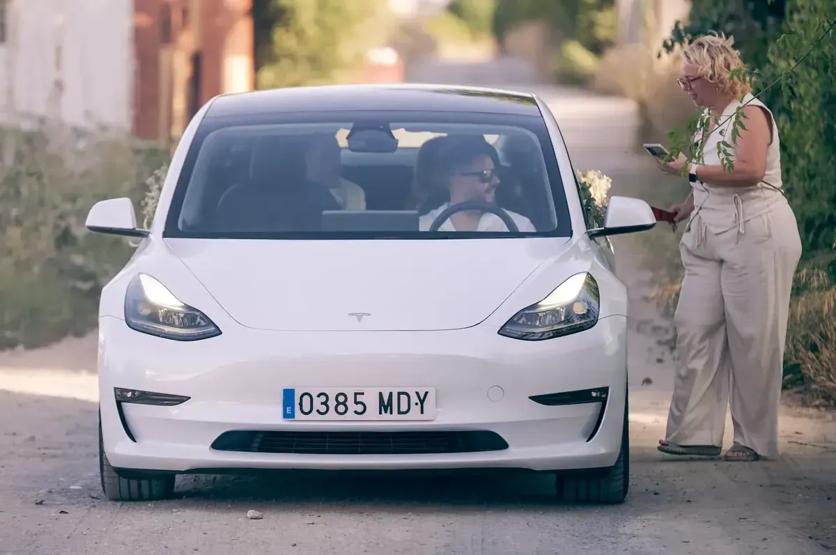 Coche blanco en una calle estrecha con una persona de pie en el lado del pasajero sosteniendo flores.