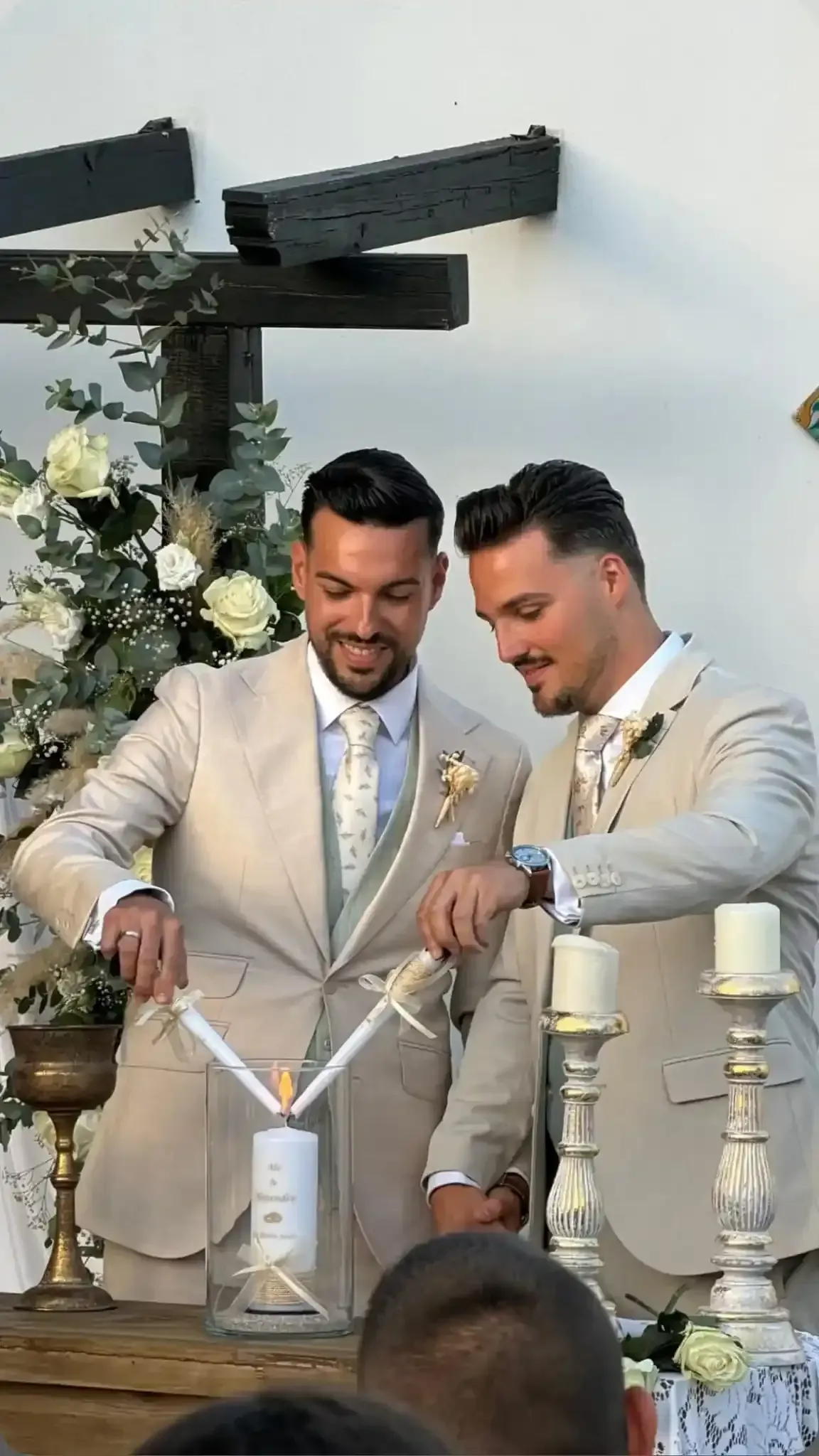 Dos hombres vestidos con trajes beige cortan un pastel blanco en una mesa de celebración decorada.