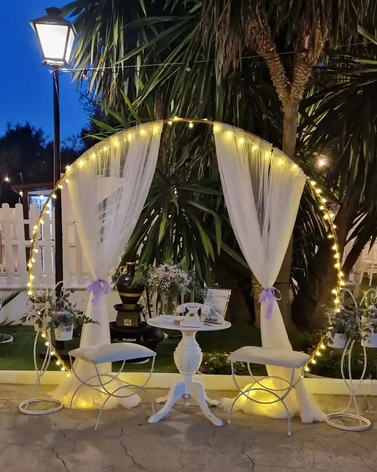 Mesa exterior decorada bajo un arco iluminado con cortinas de tela blanca y palmeras al atardecer.