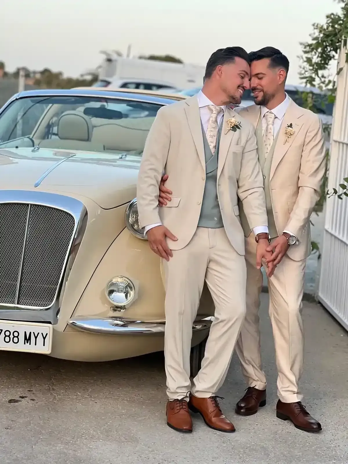 Dos hombres con trajes color crema, de pie junto a un descapotable clásico del mismo color, sonríen muy juntos al aire libre.