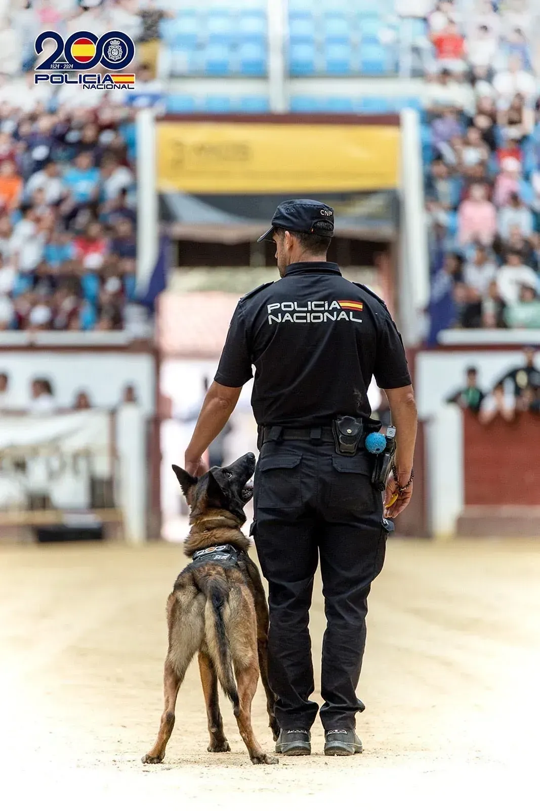Un oficial de policía de pie junto a un perro frente a una plaza de toros.