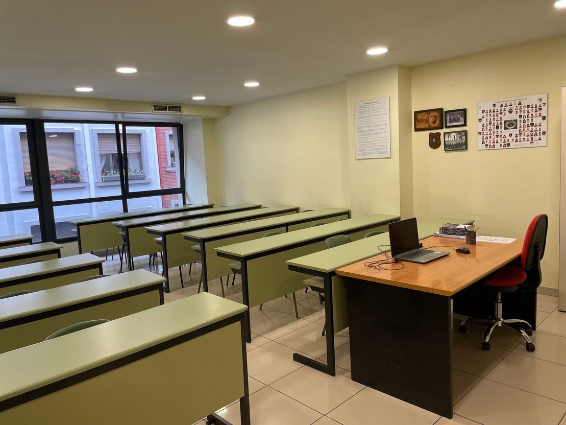 Un aula con filas de mesas y sillas y un escritorio con una computadora portátil encima.