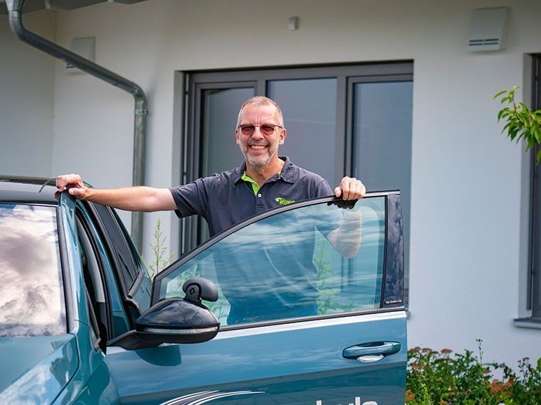 Ein Mann mit Brille lächelt, er steht neben einem türkisfarbenen Auto mit offener Tür vor einem Haus.