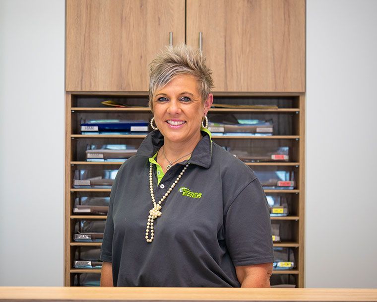 Eine Frau in einem grauen Poloshirt lächelt an einem Empfangstresen; im Hintergrund sind Aktenordner zu sehen.