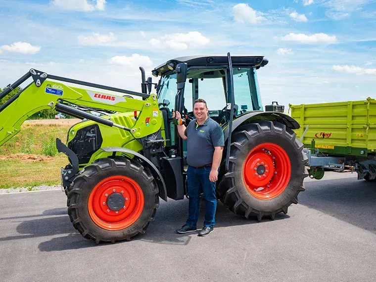 Ein Mann steht an einem sonnigen Tag im Freien neben einem grün-schwarzen Claas-Traktor mit Anhänger.