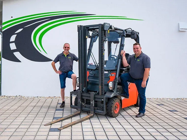 Zwei Männer posieren mit einem Gabelstapler vor einer weißen Wand mit einem Straßenlogo.