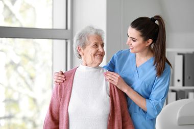 Une aide-soignante en blouse bleue aide une femme âgée à enfiler un gilet rose à l'intérieur.