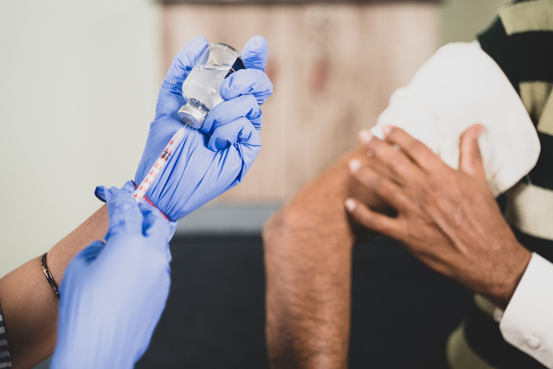Un agent de santé ganté de bleu tient une seringue à vaccin près du bras nu d'un patient masqué.