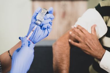 Main gantée préparant une seringue à vaccin à côté d'une personne tenant un coton sur son bras.