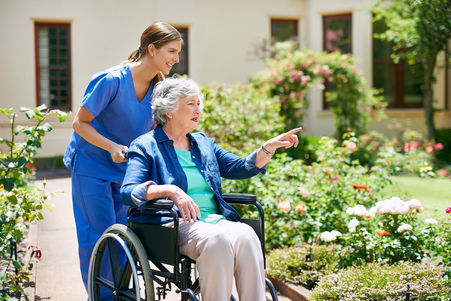 Une aide-soignante en bleu se tient à côté d'une personne en fauteuil roulant qui désigne un jardin fleuri ensoleillé.