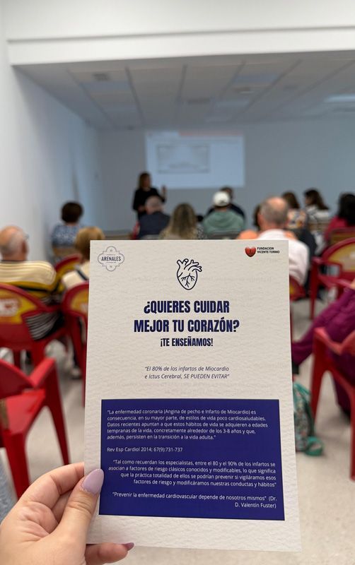 Una persona sostiene un folleto frente a un grupo de personas sentadas en sillas rojas.
