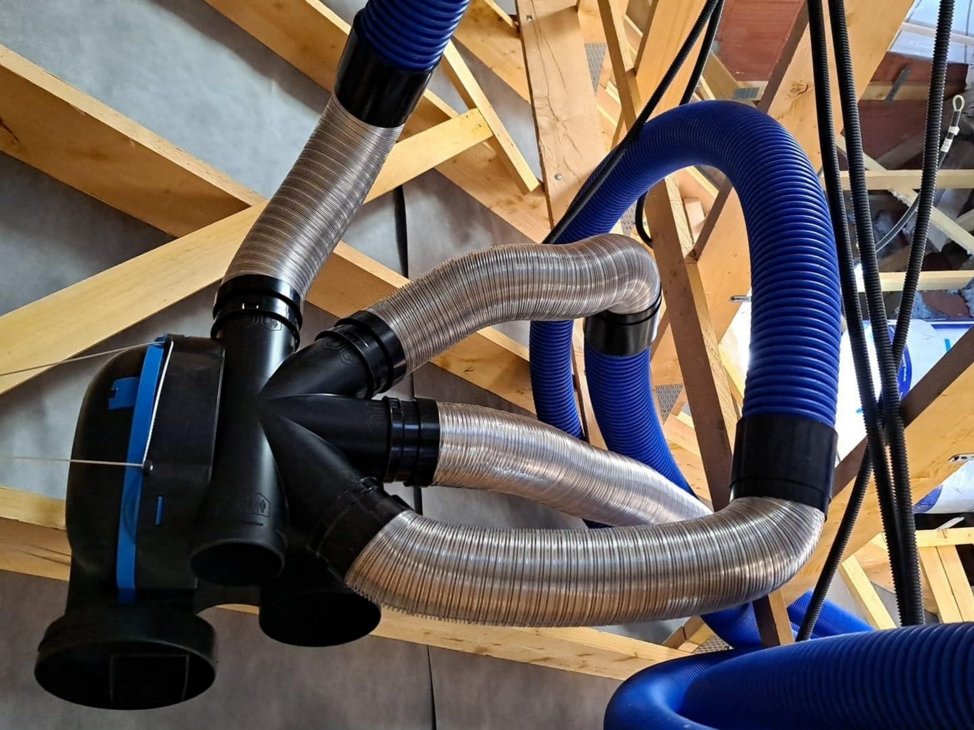 Des conduits d'aération bleus et argentés reliés à une unité de traitement d'air dans un grenier avec des poutres en bois.