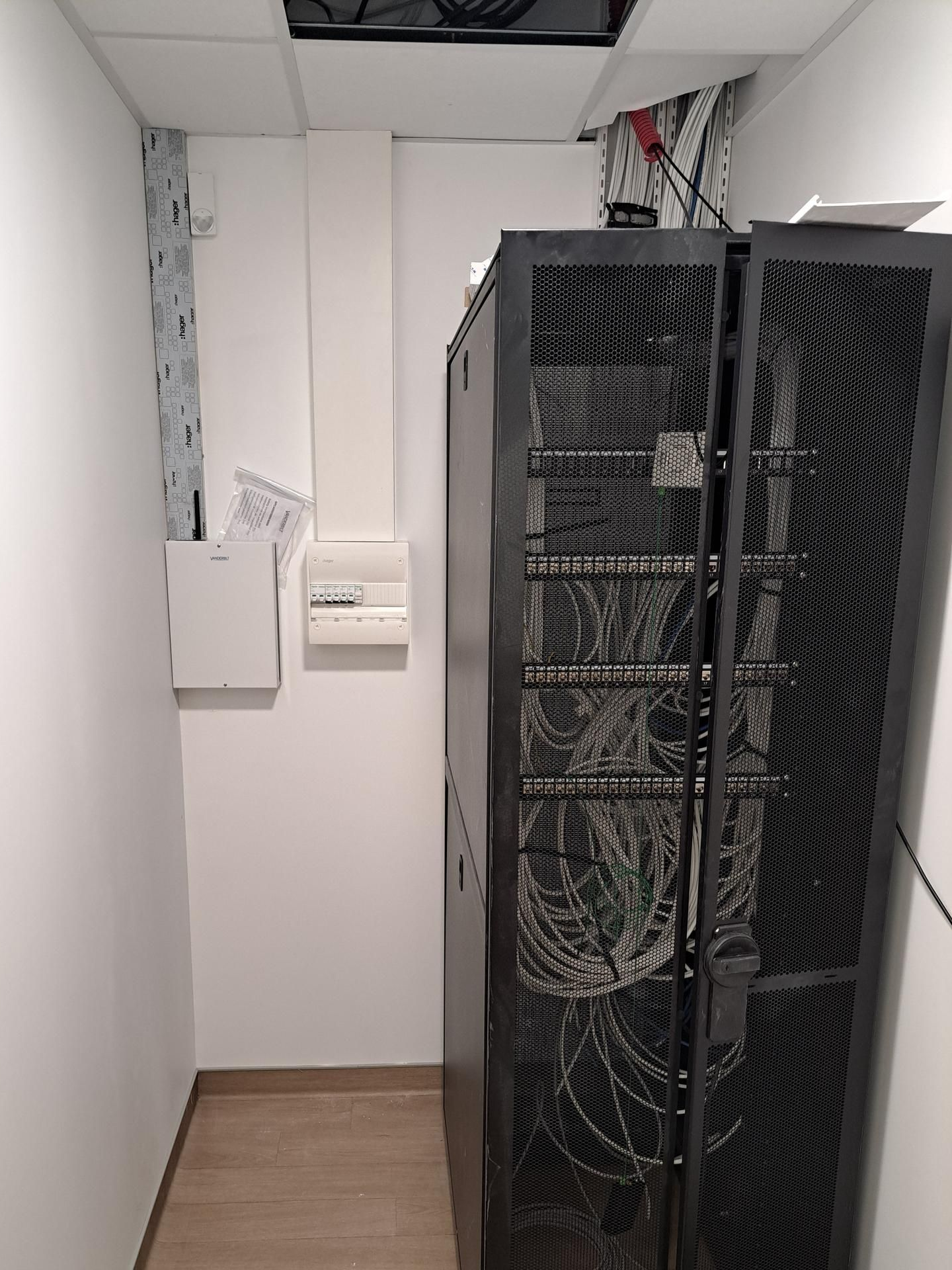Baie de serveurs avec câbles dans une petite pièce, tableau électrique, conduits et câblage apparent au plafond.