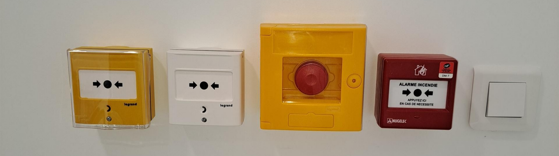 Des prises électriques murales, un bouton d'alarme incendie et un interrupteur sur un mur blanc.