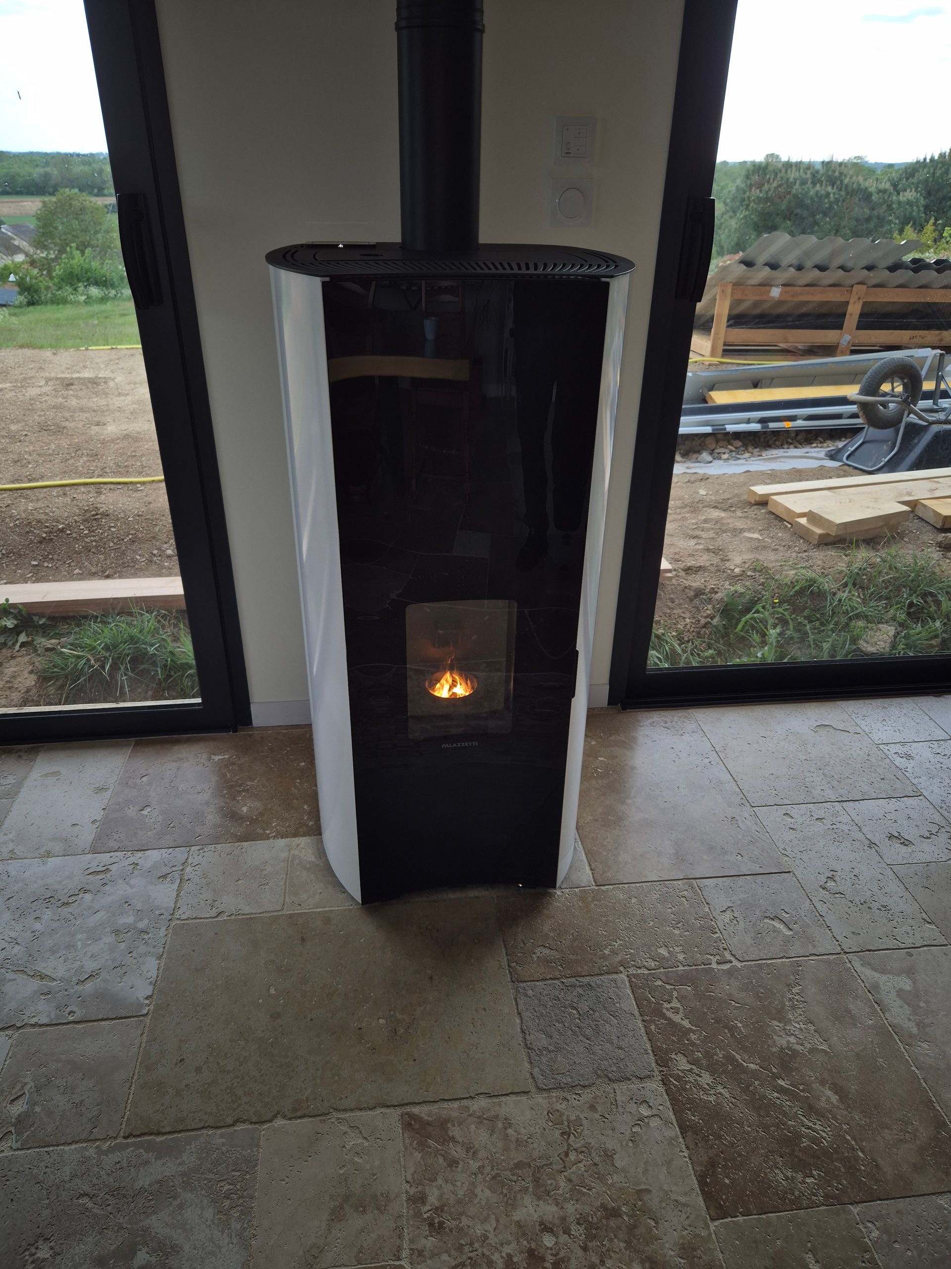 Petit poêle à bois noir et blanc avec un feu allumé, placé à l'intérieur près de grandes fenêtres.