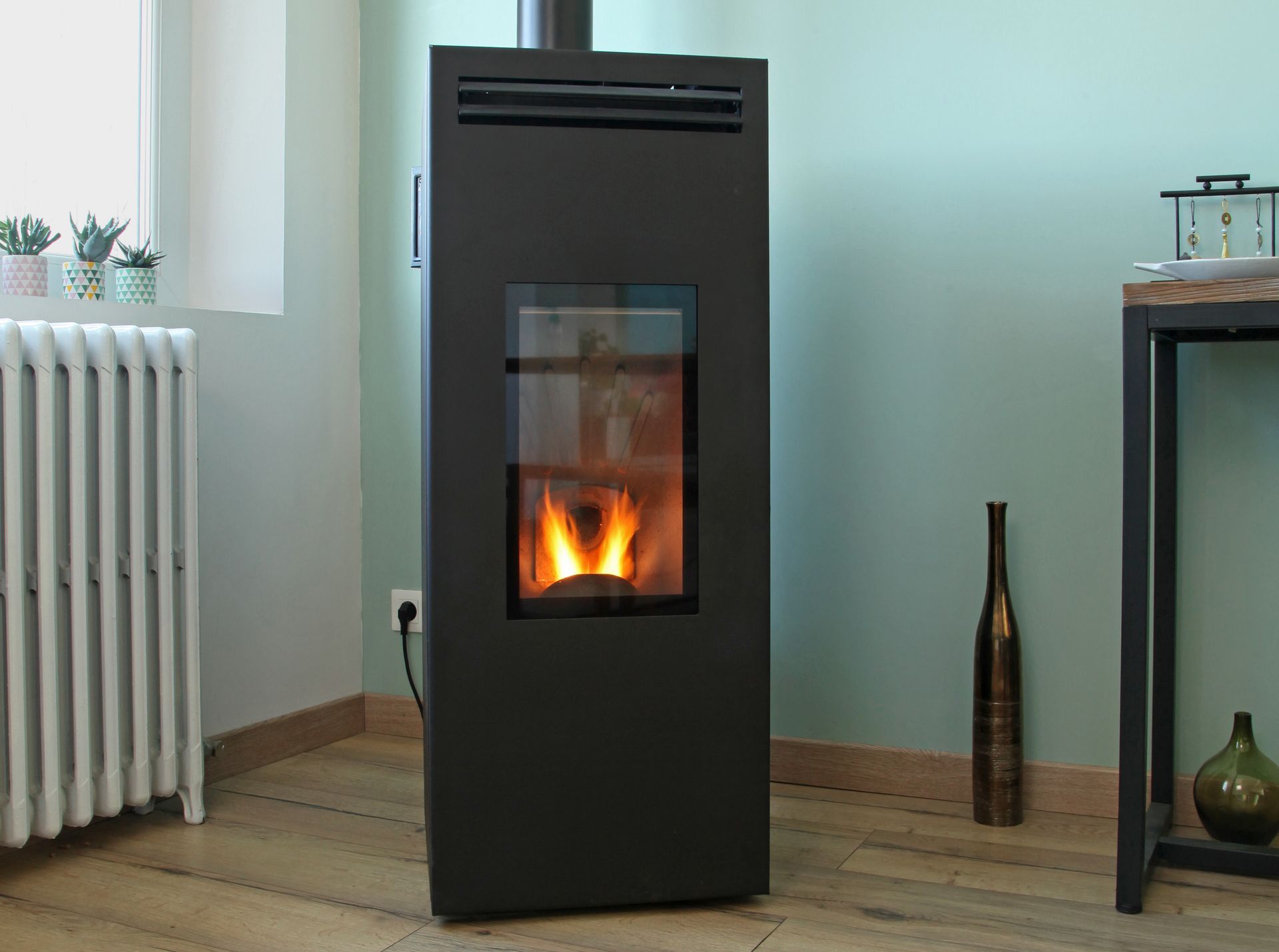 Cheminée noire autoportante avec flammes visibles dans une pièce lumineuse, à côté d'un radiateur et d'une table d'appoint.