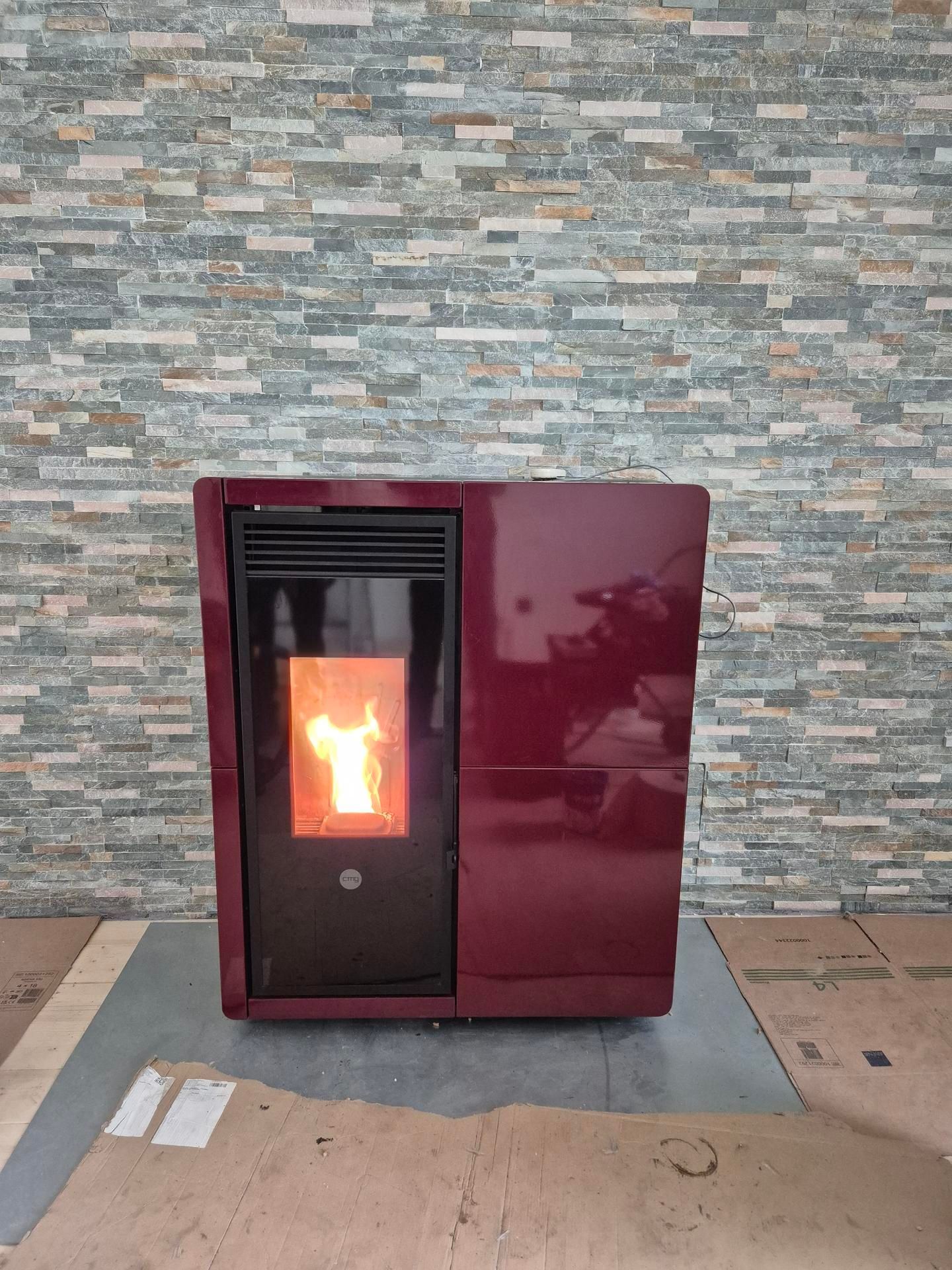 Poêle à granulés marron avec un feu incandescent, adossé à un mur de pierre sur un sol carrelé.