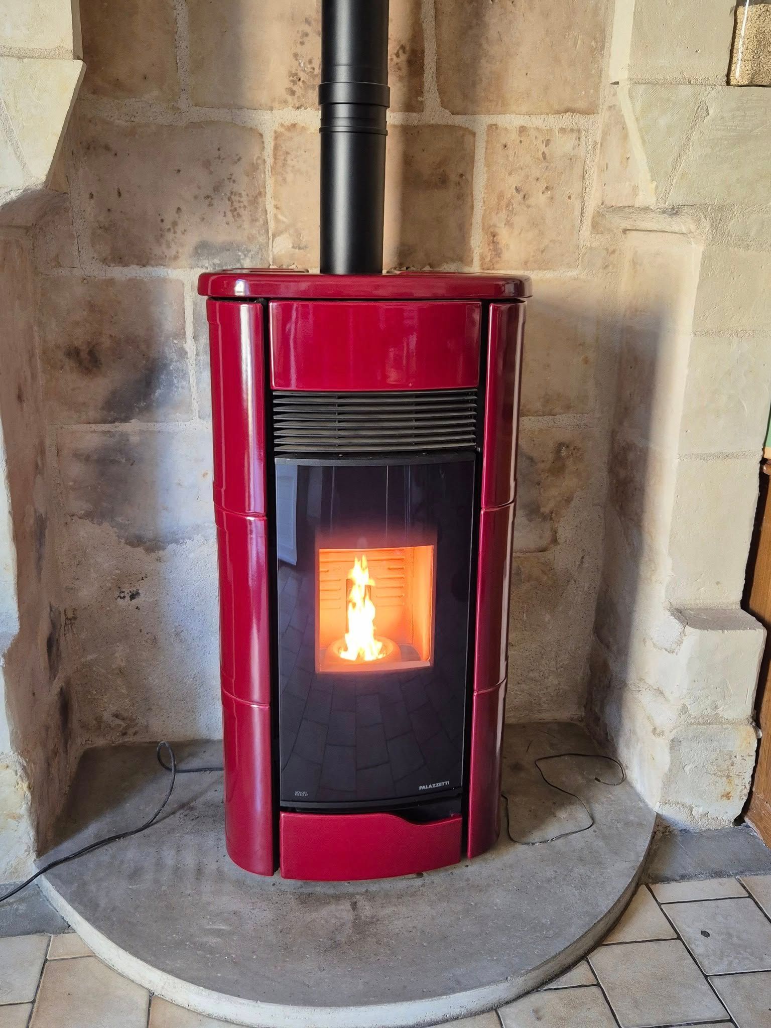 Poêle à bois rouge avec une flamme visible, installé dans un foyer en pierre contre un mur en pierre.