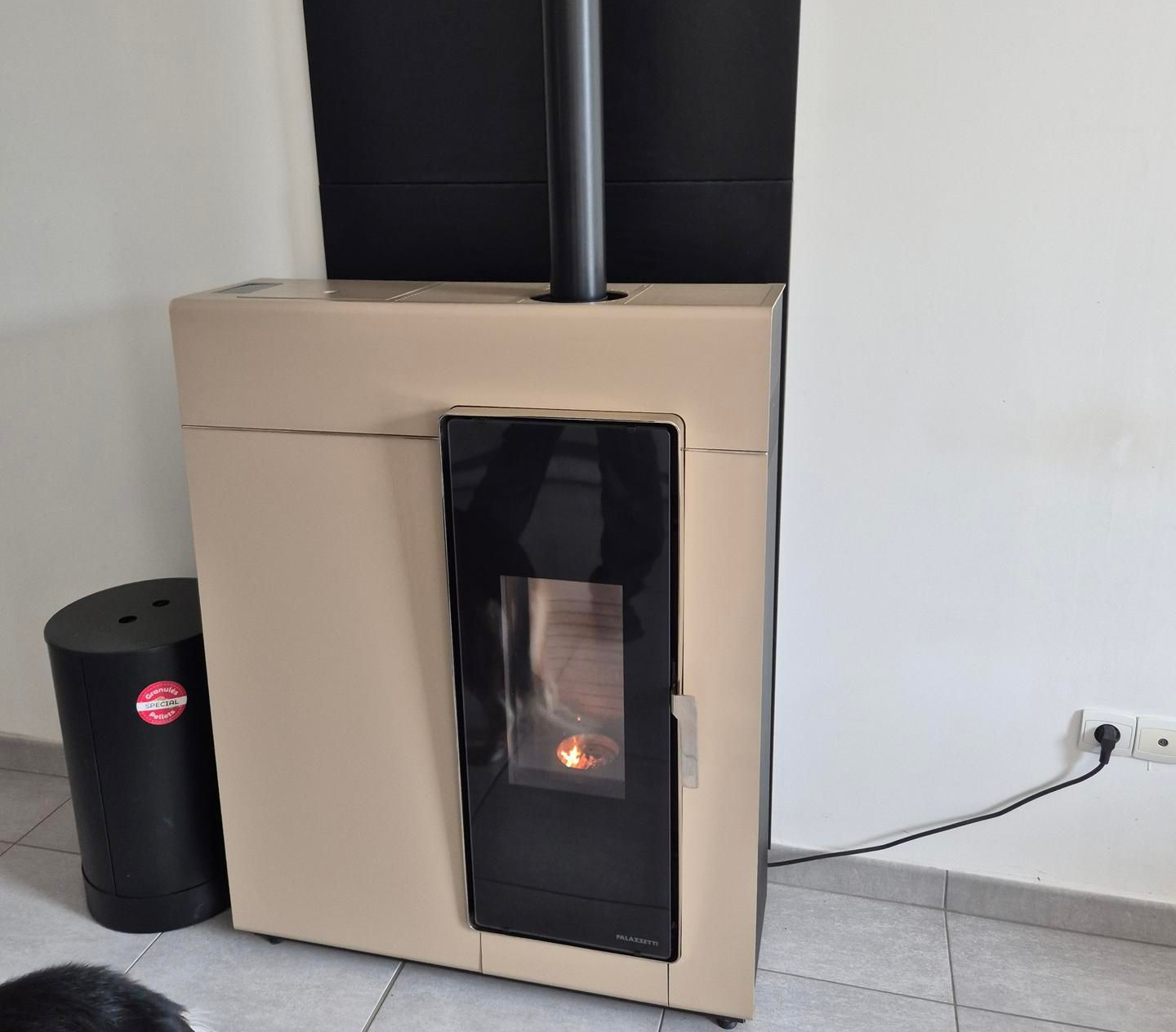 Poêle à bois beige avec porte vitrée noire et conduit de cheminée, à côté d'une poubelle noire sur un sol carrelé.