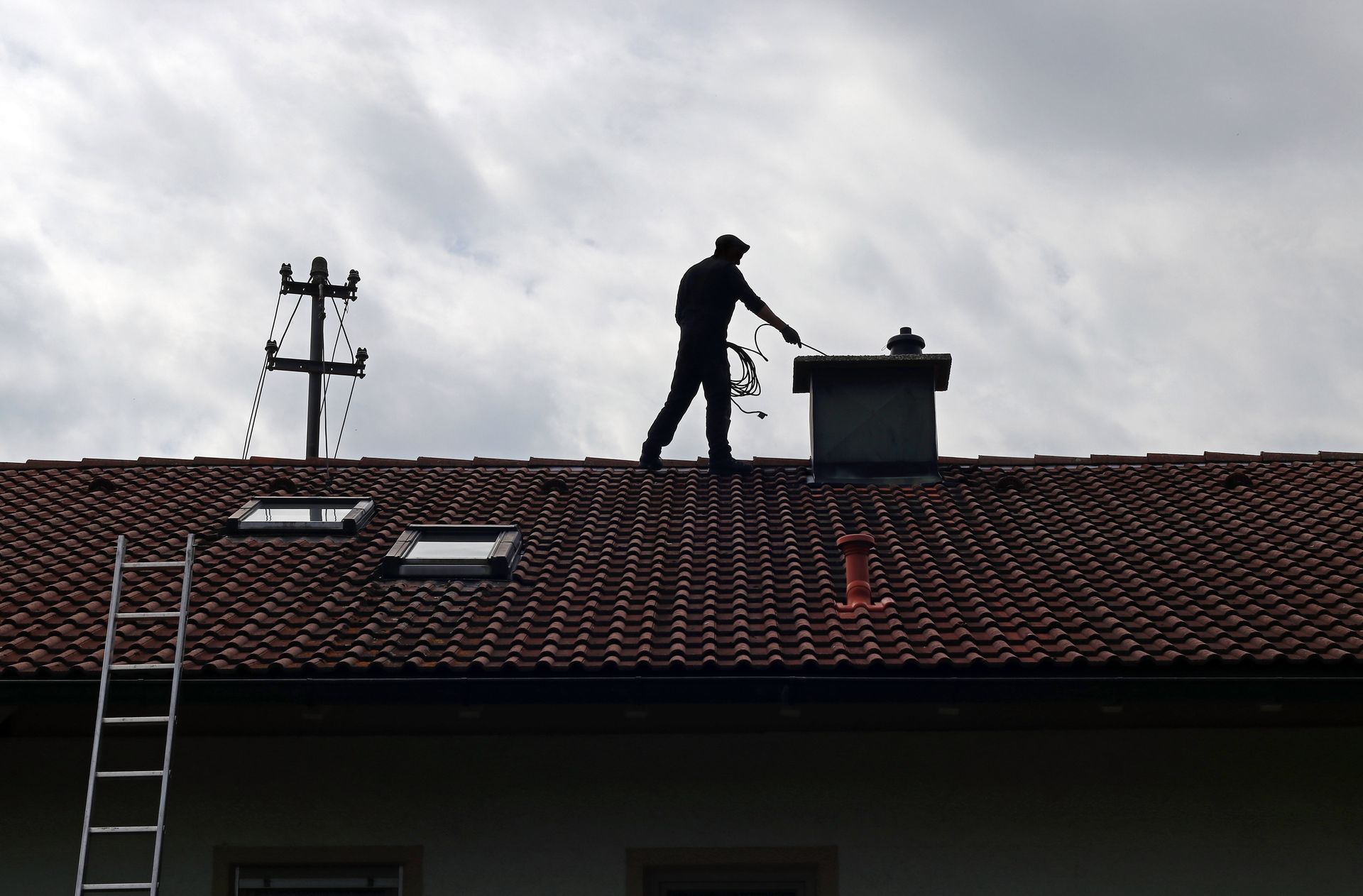 Silhouette d'un couvreur sur un toit de tuiles, avec une échelle et un poteau électrique, sous un ciel nuageux.