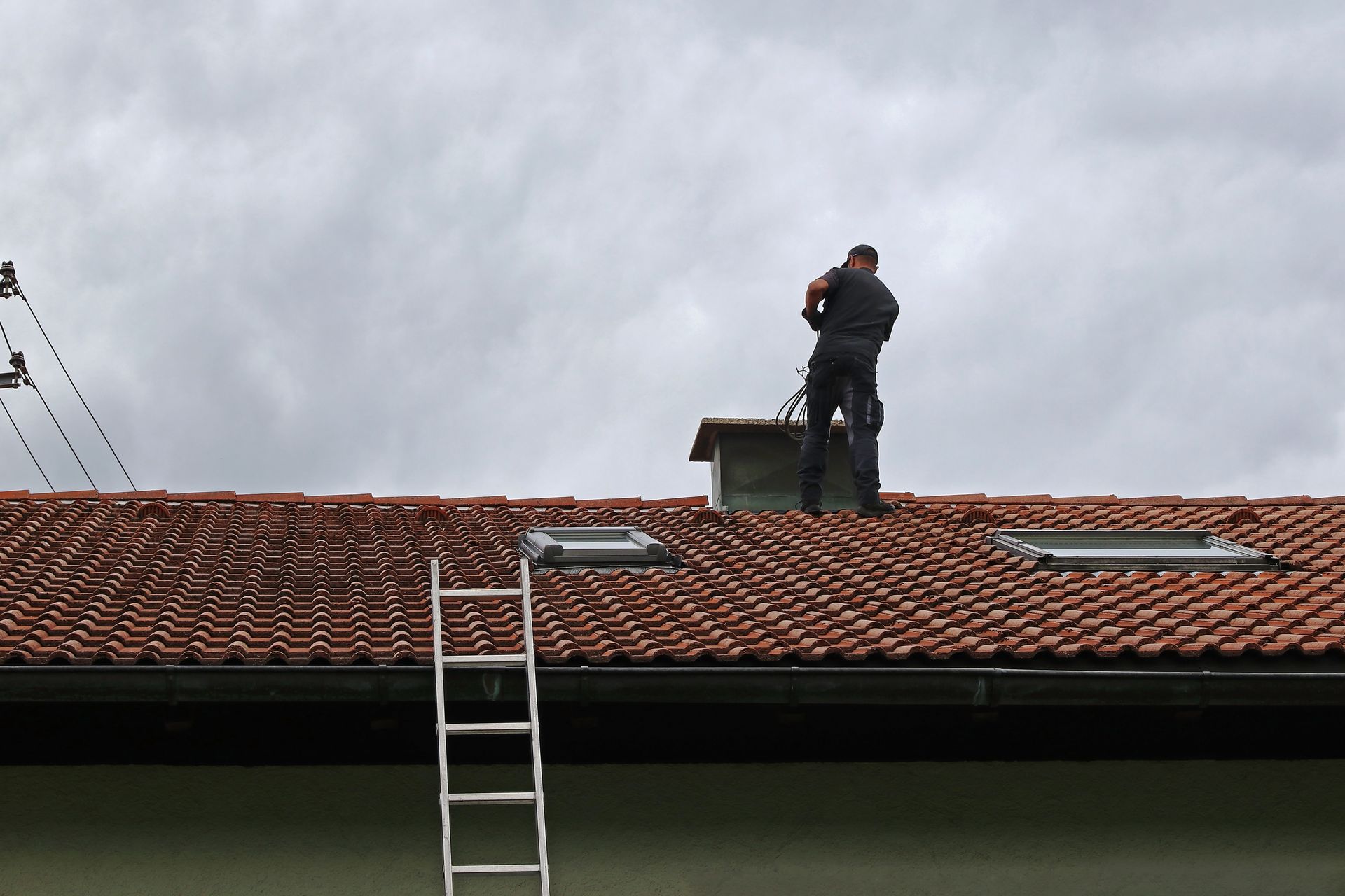 Ouvrier sur un toit de tuiles rouges, à côté d'une échelle, sous un ciel nuageux.