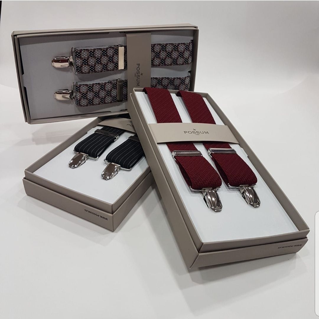 Tres tirantes en caja de color gris, burdeos y negro con clips plateados exhibidos sobre una superficie blanca.