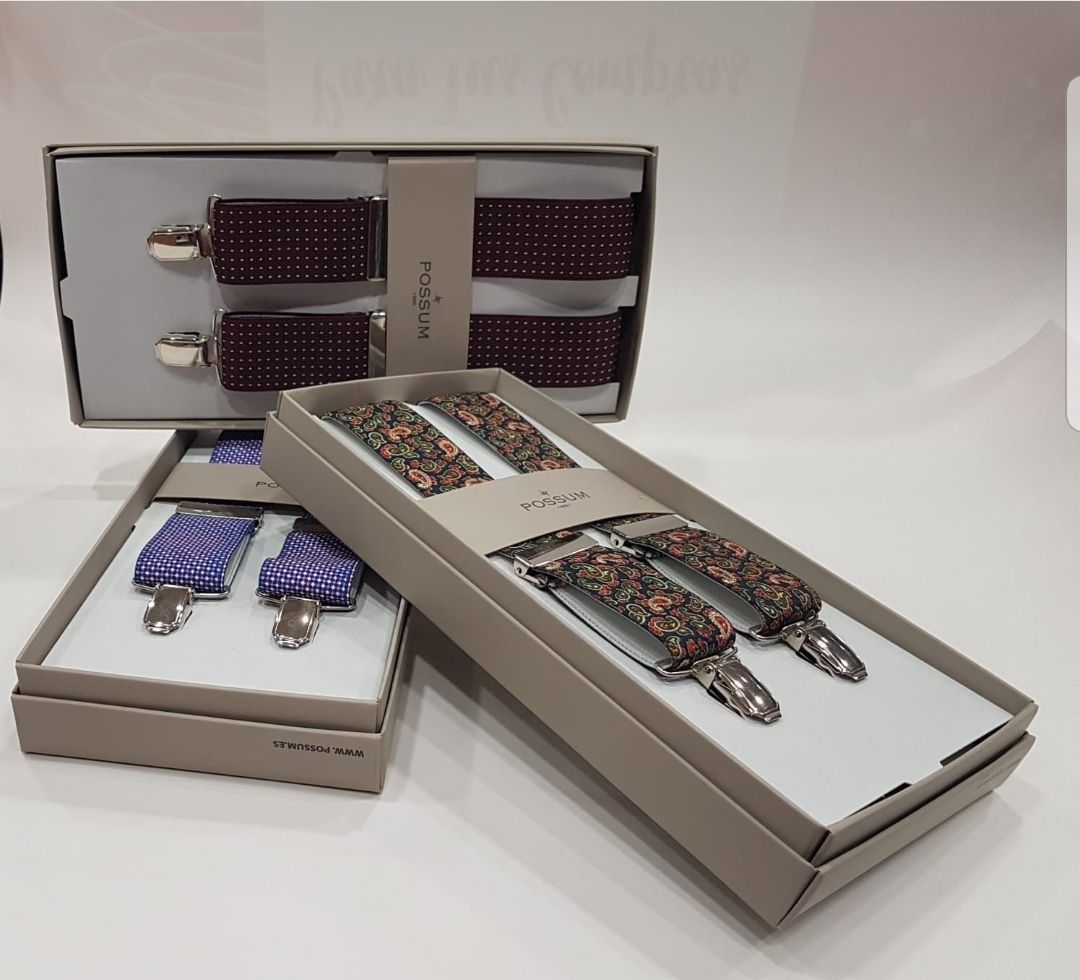 Tres pares de tirantes para hombre en cajas abiertas: burdeos, florales y morados a cuadros, sobre una superficie blanca.