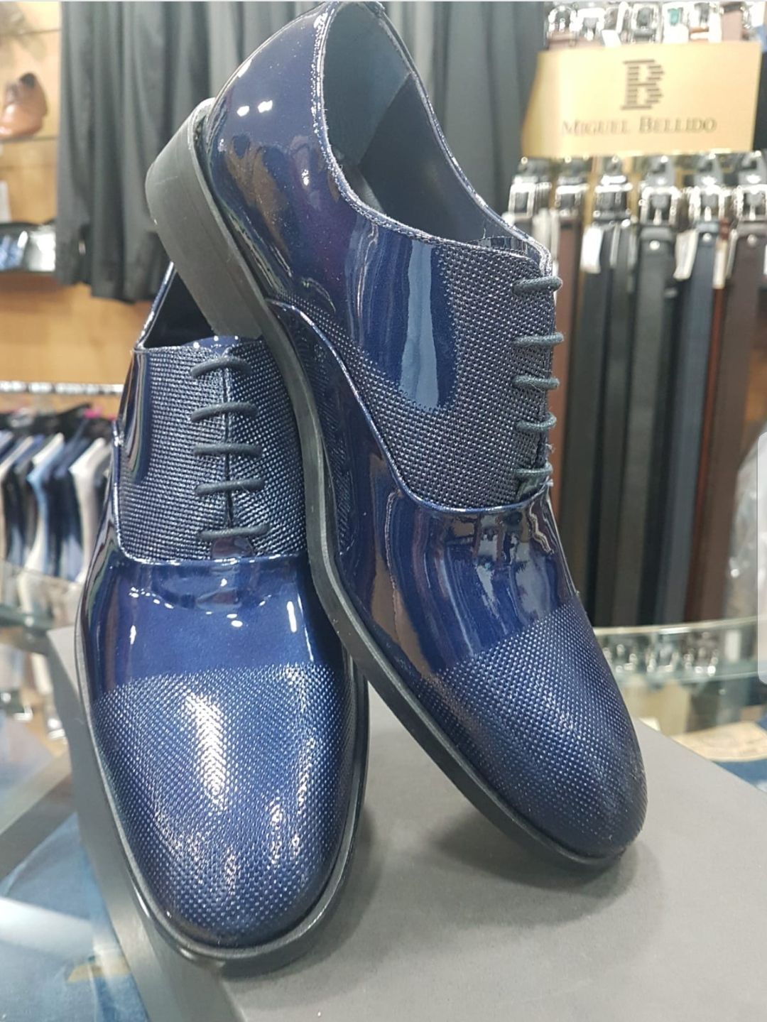 Zapatos de vestir para hombre de piel charol azul con puntera texturizada y detalle de cordones, expuestos en una superficie minorista.