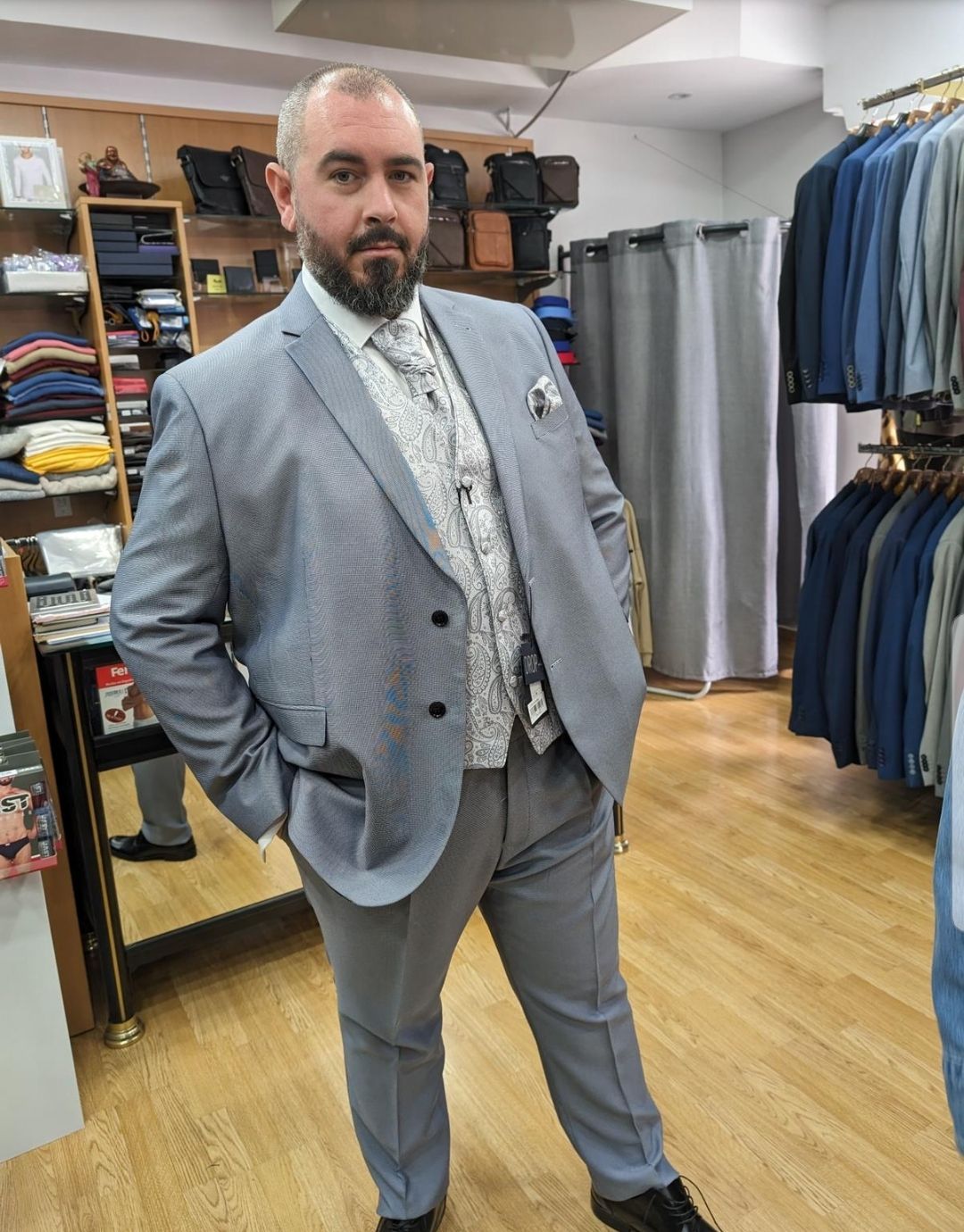 Un hombre con traje gris, chaleco y corbata estampada, con las manos en los bolsillos, está en una tienda de ropa.