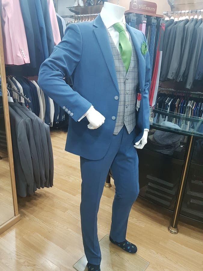 Maniquí con traje azul, chaleco gris, corbata verde y pañuelo de bolsillo, de pie en una tienda de ropa.