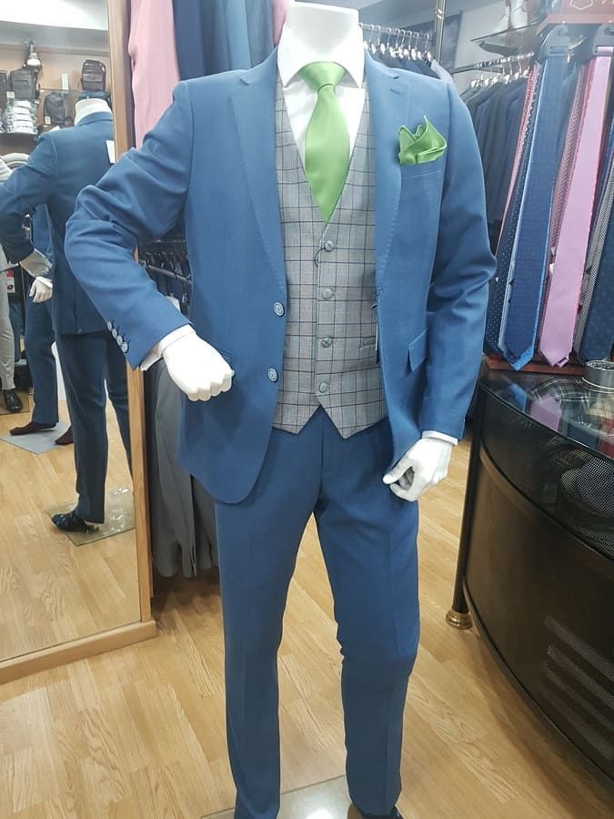 Maniquí con traje azul, chaleco gris, corbata verde y pañuelo de bolsillo en una tienda de ropa.
