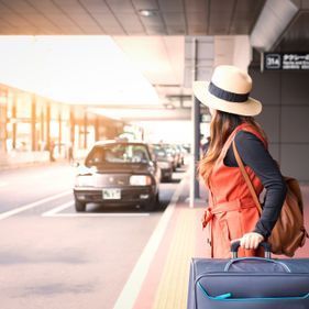 Une femme en manteau et chapeau orange attend un taxi avec une valise devant un immeuble.