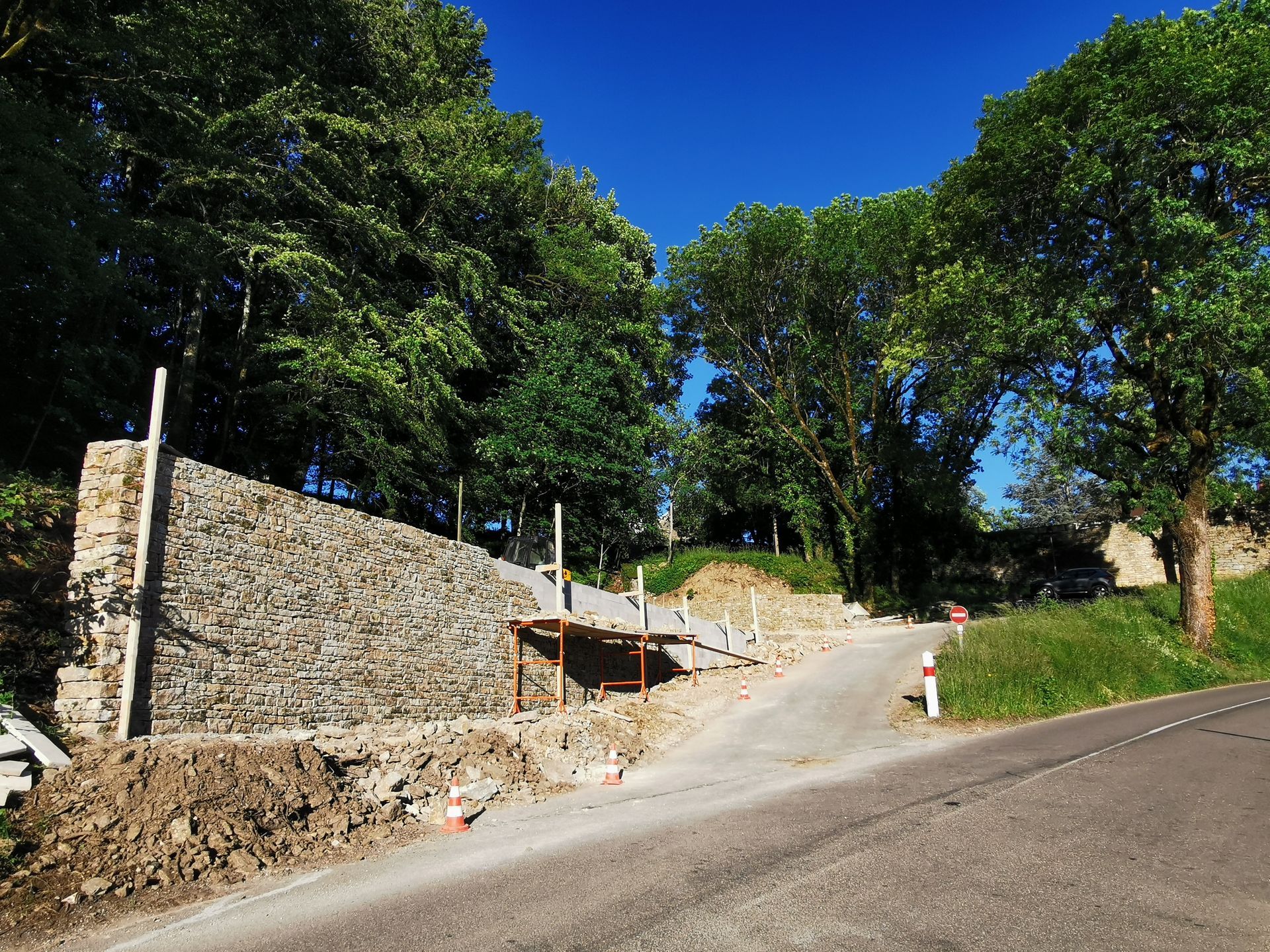 Une route avec un mur de soutènement à gauche, des arbres et des travaux en cours.