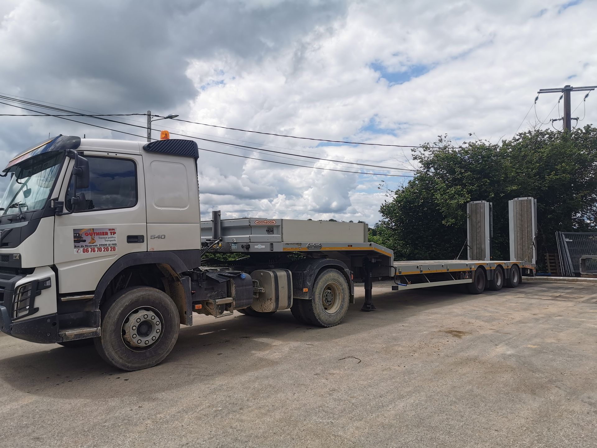 Camion blanc avec remorque plateau transportant de hauts piliers en béton, stationné sur du gravier.
