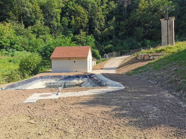 Petit bâtiment beige avec toit en tuiles rouges, zone bétonnée et une tour.