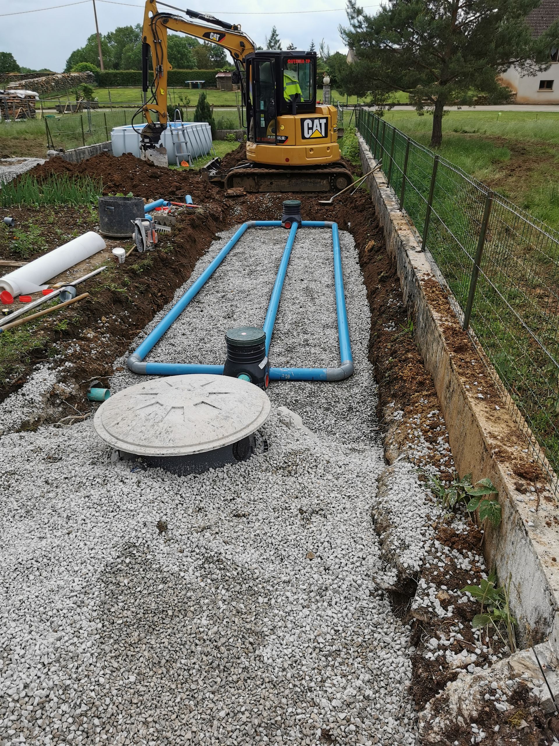 Chantier : excavatrice, lit de gravier, tuyaux bleus et une fosse pour système septique.