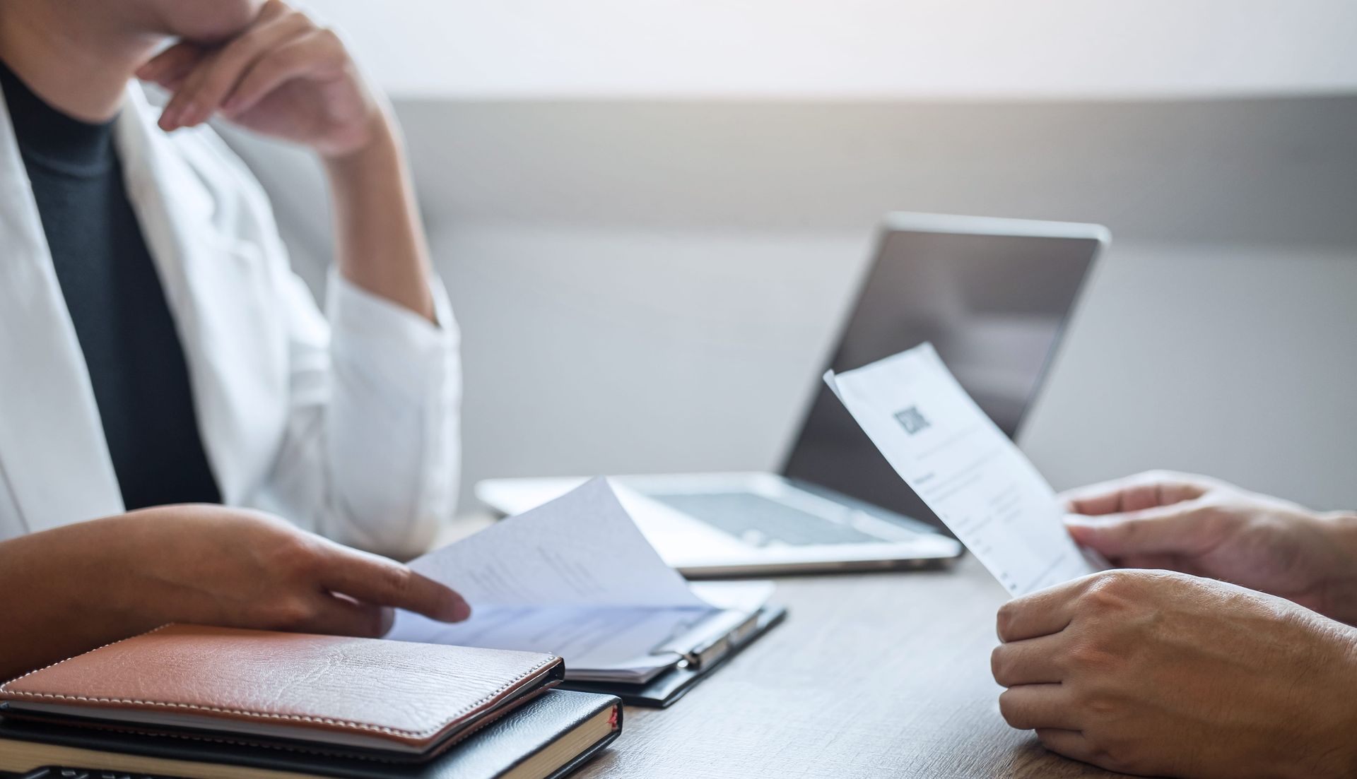 Two people exchanging documents across a desk with a laptop and notebook nearby