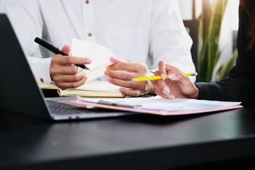 Dos personas revisando notas en un escritorio con una computadora portátil, bolígrafos y documentos.