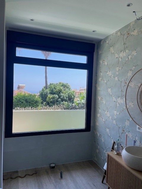 Habitación pequeña con una ventana grande, papel pintado floral, lavabo con tocador y vistas a los árboles del exterior.
