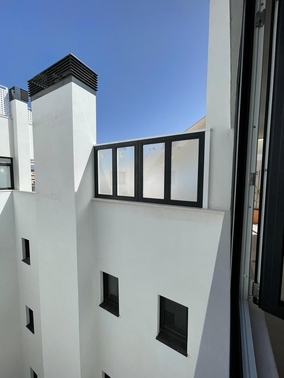 Edificio de fachada blanca con marcos de ventanas negros y barandilla en la azotea bajo un cielo azul.