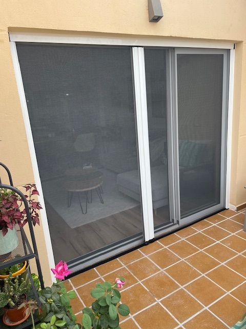 Puerta corrediza de cristal con mosquitera gris, que da a un patio con baldosas de terracota y plantas en macetas.