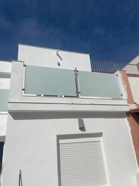 Fachada blanca del edificio con barandilla de cristal en la azotea y persiana cerrada bajo un cielo azul.