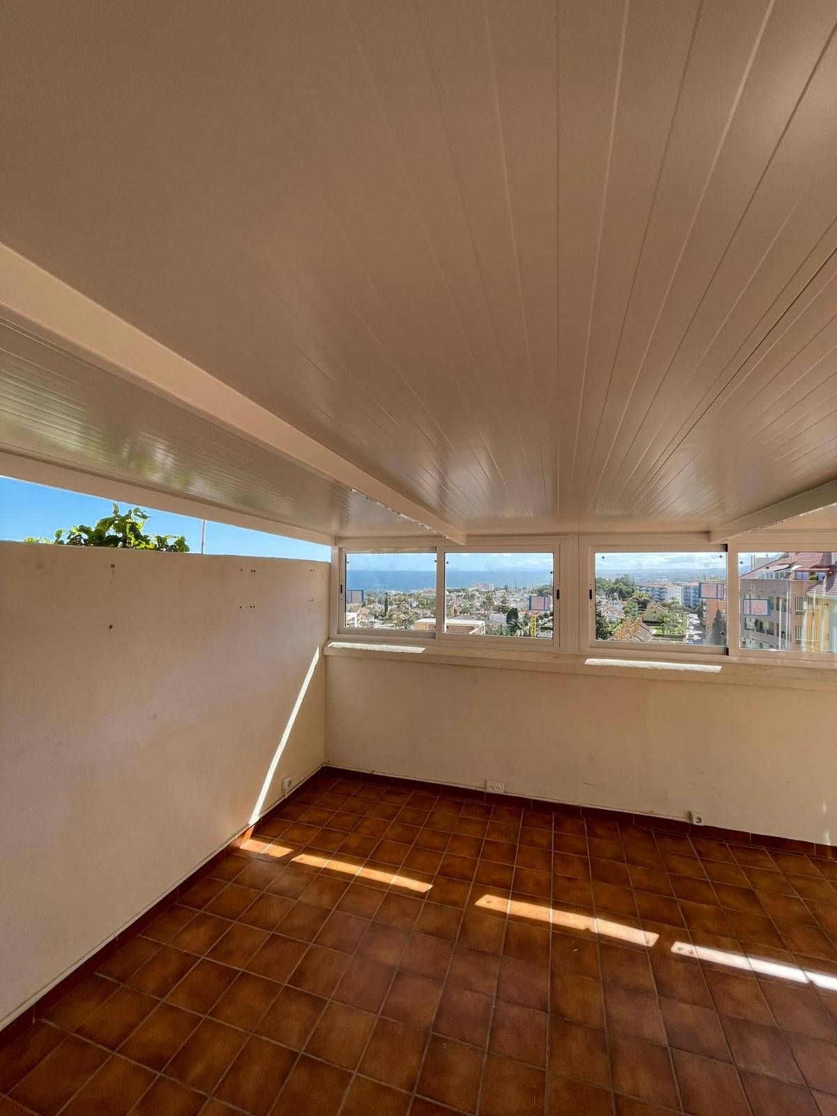 Balcón cubierto vacío con suelo de baldosas de terracota, paredes blancas y vistas a la ciudad a través de ventanas estrechas.