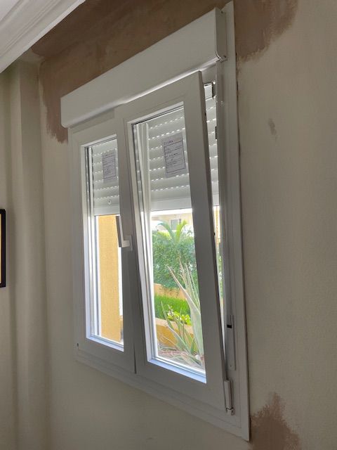 Ventana blanca en una pared beige con un mosaico visible alrededor del marco, que deja ver vegetación en el exterior.